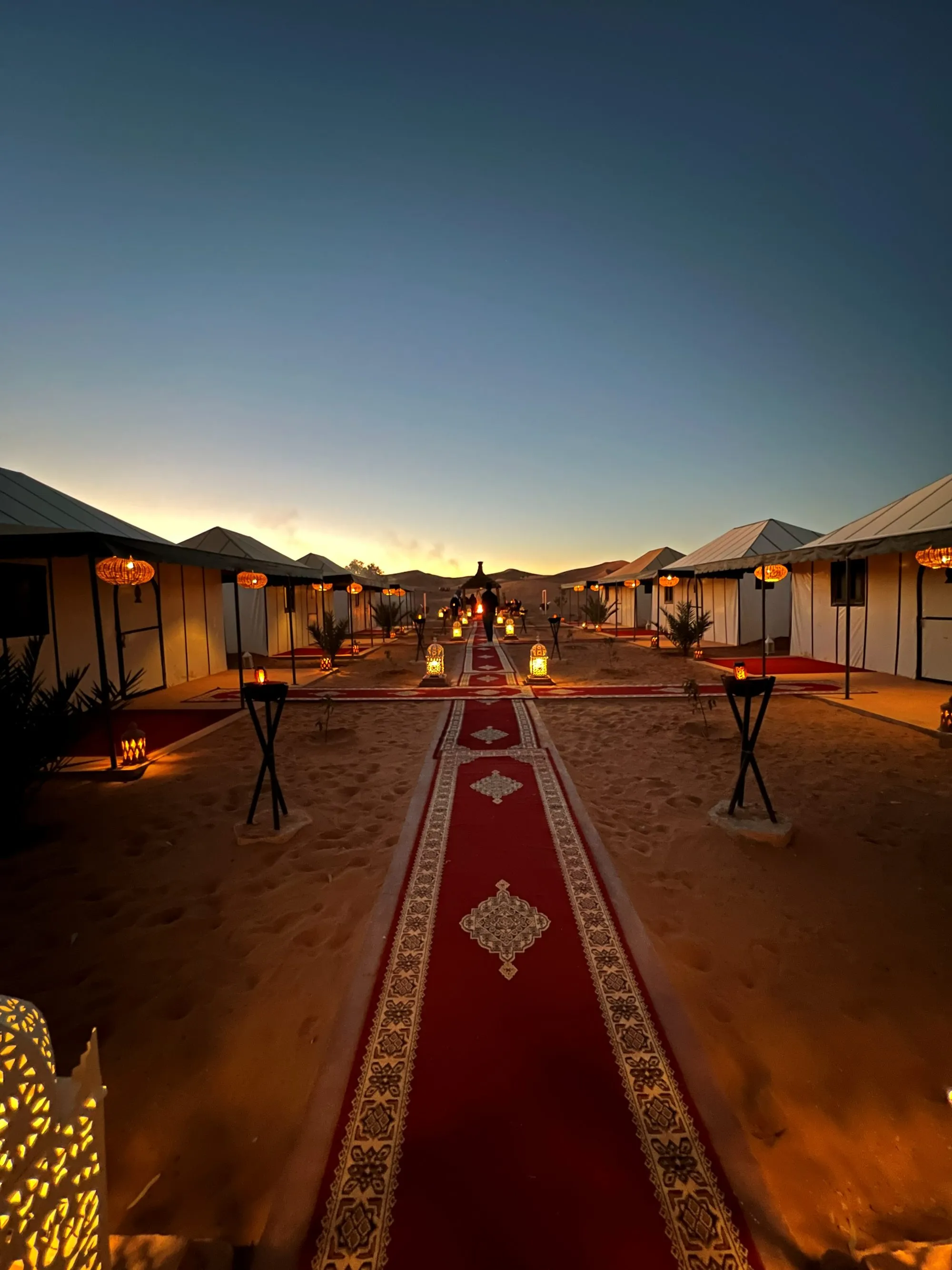 A desert camp at dusk with a carpeted path across the sand
